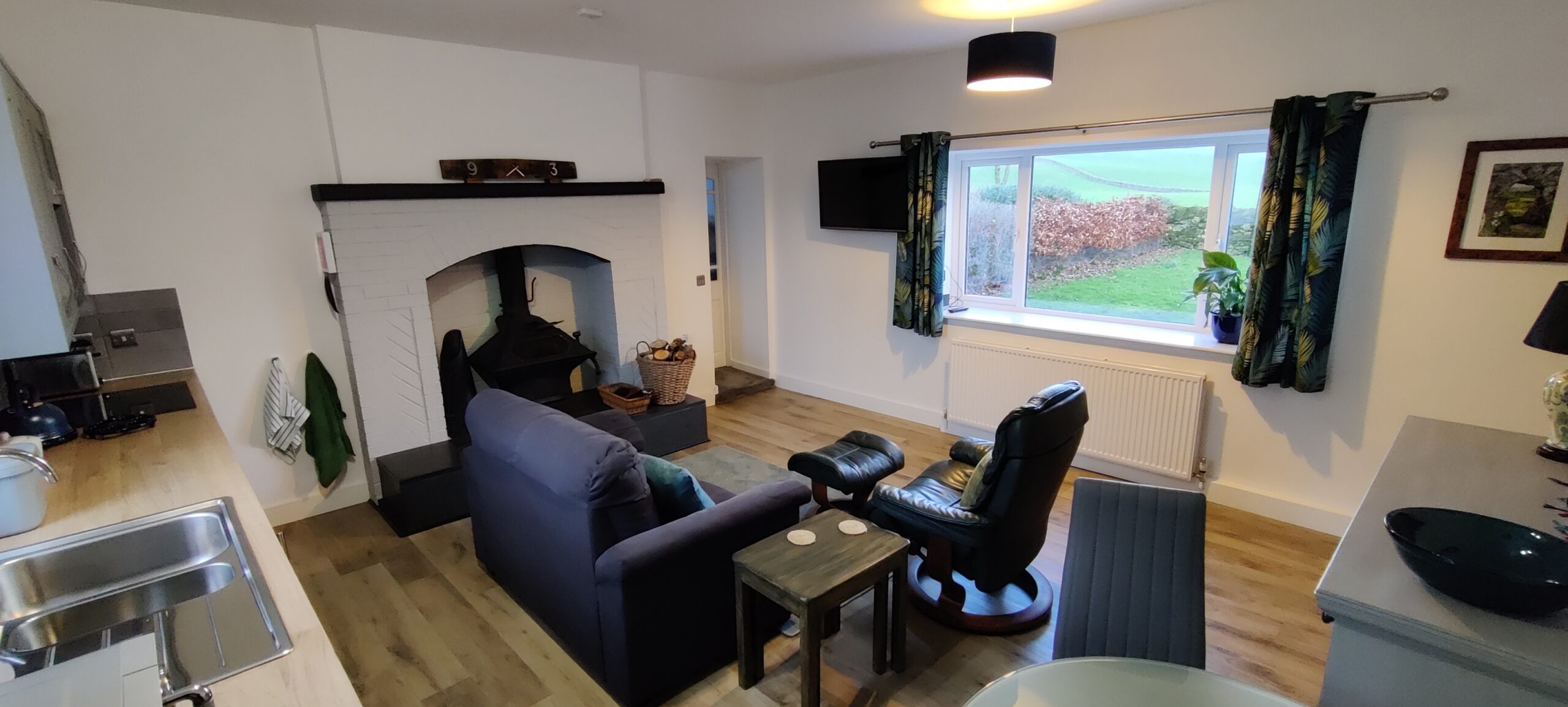 Kitchen Lounge, view from kitchen corner. Shows log burner, sofa and comfy chair, and TV.